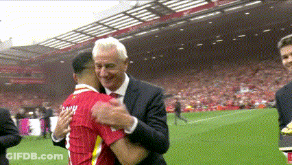 A Man In A Suit And Tie Is Hugging A Man In A Red Shirt On A Soccer Field GIF