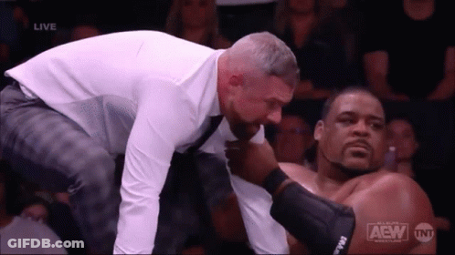 A Man In A Suit And Tie Is Kneeling Down Next To A Wrestler With The Word Aew On The Bottom Of The Screen GIF
