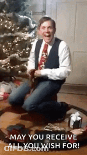 A Man In A Suit And Tie Is Kneeling In Front Of A Christmas Tree And A Train Set . GIF
