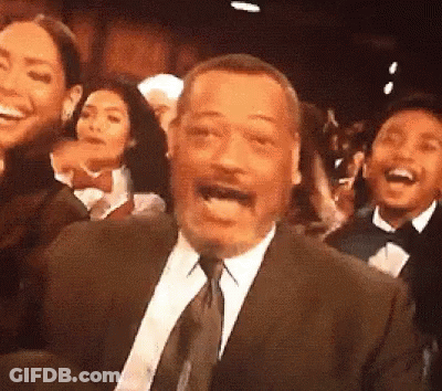 A Man In A Suit And Tie Is Laughing In Front Of A Crowd GIF