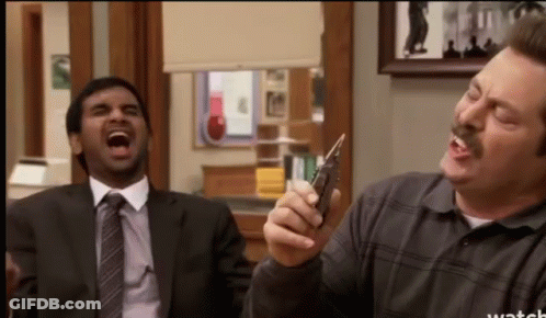 A Man In A Suit And Tie Is Laughing While Another Man Holds A Cell Phone GIF