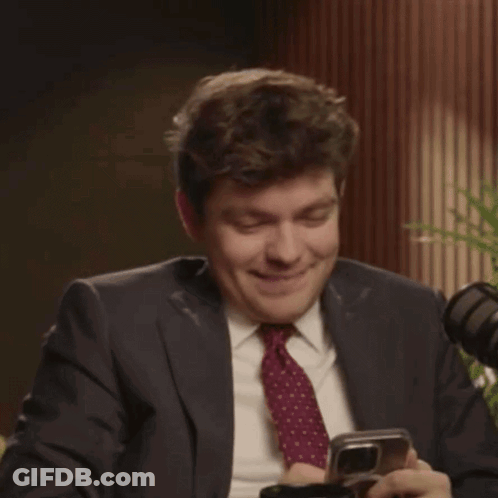 A Man In A Suit And Tie Is Laughing While Holding A Phone GIF