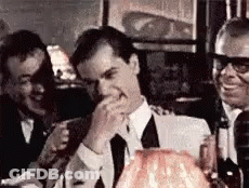 A Man In A Suit And Tie Is Laughing While Sitting At A Table With Two Other Men . GIF