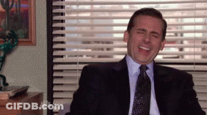 A Man In A Suit And Tie Is Laughing While Sitting In Front Of A Window With Blinds . GIF