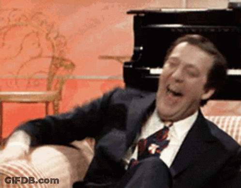 A Man In A Suit And Tie Is Laughing While Sitting On A Couch In Front Of A Piano GIF