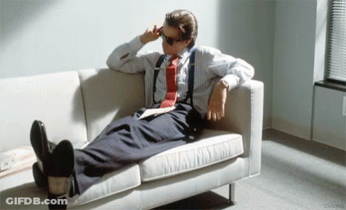 A Man In A Suit And Tie Is Laying On A White Couch With His Feet Up . GIF