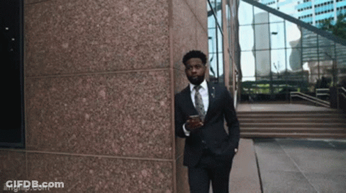 A Man In A Suit And Tie Is Leaning Against A Building While Holding A Cup Of Coffee GIF