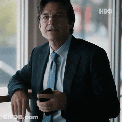 A Man In A Suit And Tie Is Leaning Against A Railing Holding A Cell Phone With The Hbo Logo In The Background GIF