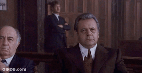 A Man In A Suit And Tie Is Looking At The Camera While Another Man Stands Behind Him . GIF
