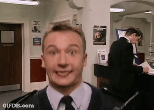 A Man In A Suit And Tie Is Making A Funny Face While Another Man Stands Behind Him . GIF