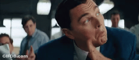 A Man In A Suit And Tie Is Making A Funny Face While Sitting In A Room With Other Men . GIF