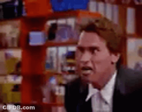 A Man In A Suit And Tie Is Making A Funny Face While Standing In Front Of A Bookshelf . GIF