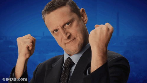 A Man In A Suit And Tie Is Making A Funny Face With His Fist In The Air GIF