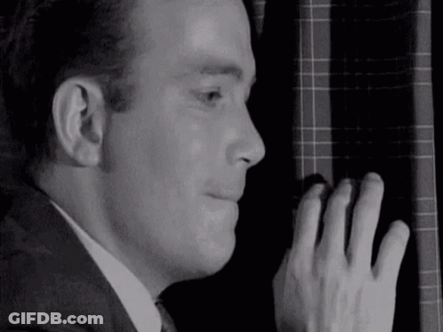 A Man In A Suit And Tie Is Peeking Out Of A Window With His Hand On The Curtain . GIF