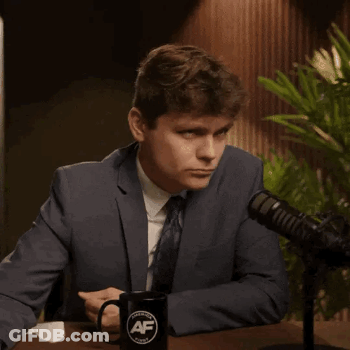 A Man In A Suit And Tie Is Pointing At A Mug That Says Af GIF
