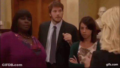 A Man In A Suit And Tie Is Pointing At A Woman In A Purple Sweater . GIF