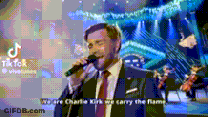 A Man In A Suit And Tie Is Singing Into A Microphone While Standing On A Stage . GIF