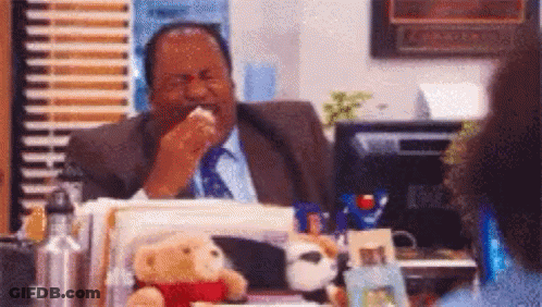 A Man In A Suit And Tie Is Sitting At A Desk And Eating A Sandwich . GIF