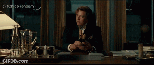 A Man In A Suit And Tie Is Sitting At A Desk And Looking At His Phone GIF
