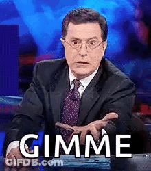 A Man In A Suit And Tie Is Sitting At A Desk And Making A Funny Face While Saying Gimme . GIF