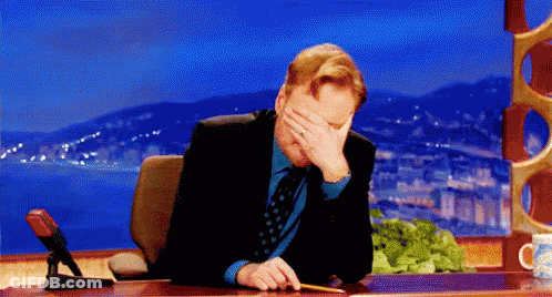 A Man In A Suit And Tie Is Sitting At A Desk Covering His Face With His Hand . GIF