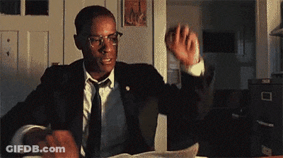 A Man In A Suit And Tie Is Sitting At A Desk Holding A Piece Of Paper . GIF