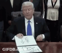 A Man In A Suit And Tie Is Sitting At A Desk Holding A Piece Of Paper GIF