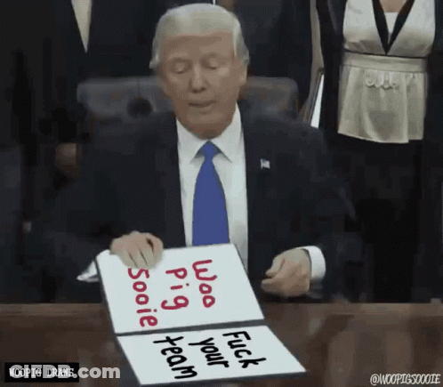 A Man In A Suit And Tie Is Sitting At A Desk Holding A Sign That Says " Woo Pig Sooie Fuck Your Team " GIF