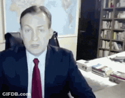 A Man In A Suit And Tie Is Sitting At A Desk In A Library . GIF
