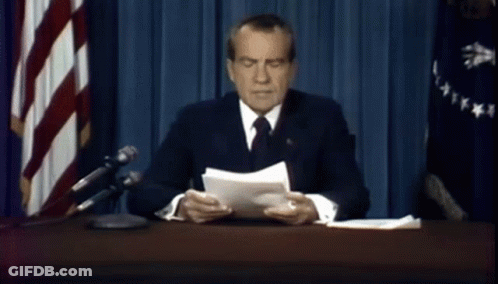 A Man In A Suit And Tie Is Sitting At A Desk In Front Of A Flag GIF