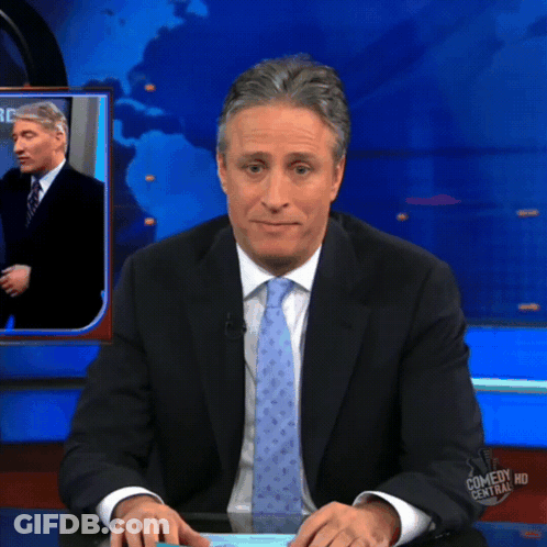 A Man In A Suit And Tie Is Sitting At A Desk In Front Of A Screen That Says Comedy Central Hd GIF