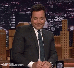 A Man In A Suit And Tie Is Sitting At A Desk In Front Of A Sign That Says Fallon Tonight GIF