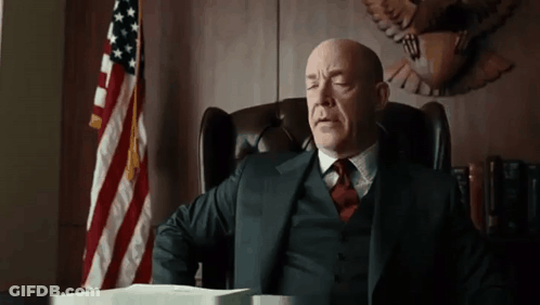 A Man In A Suit And Tie Is Sitting At A Desk In Front Of An American Flag . GIF