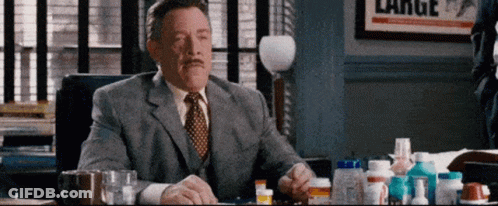 A Man In A Suit And Tie Is Sitting At A Desk With A Bunch Of Pills On It . GIF