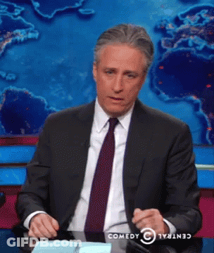 A Man In A Suit And Tie Is Sitting At A Desk With A Comedy Central Logo Behind Him GIF