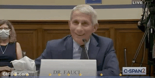 A Man In A Suit And Tie Is Sitting At A Desk With A Sign That Says Dr. Fauci GIF