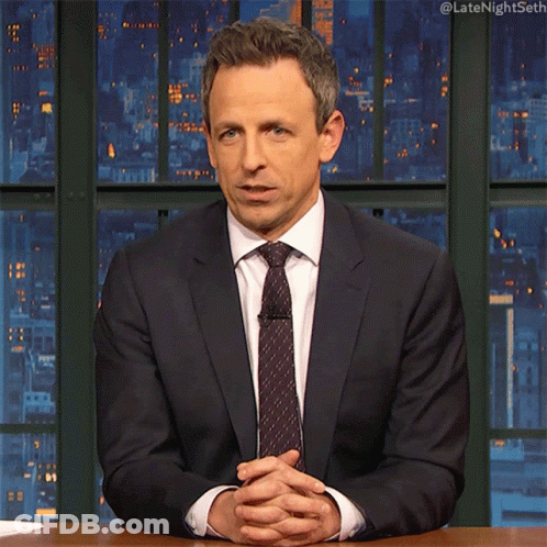 A Man In A Suit And Tie Is Sitting At A Desk With His Hands Folded GIF