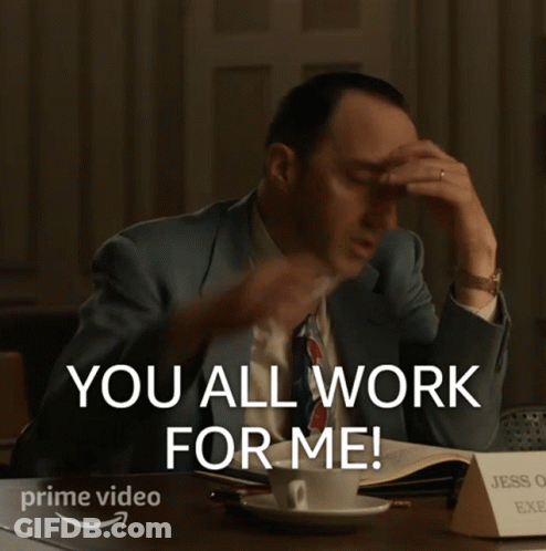 A Man In A Suit And Tie Is Sitting At A Desk With His Hands In The Air And Says You All Work For Me GIF