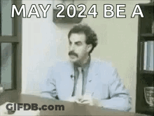 A Man In A Suit And Tie Is Sitting At A Desk With The Words `` May 2024 Be A '' Written On It . GIF