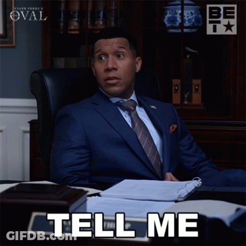 A Man In A Suit And Tie Is Sitting At A Desk With The Words Tell Me Below Him GIF
