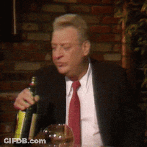 A Man In A Suit And Tie Is Sitting At A Table Holding A Bottle Of Wine GIF