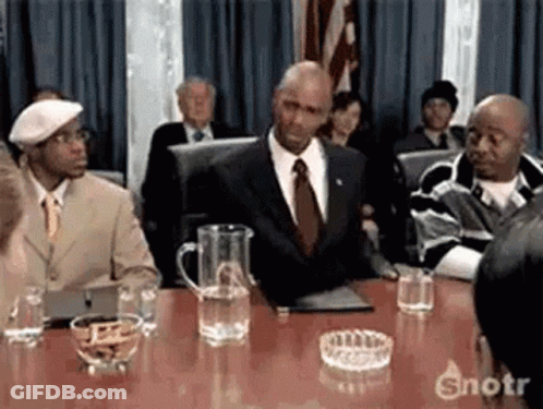 A Man In A Suit And Tie Is Sitting At A Table In Front Of A Crowd . GIF