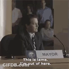 A Man In A Suit And Tie Is Sitting At A Table In Front Of A Microphone And A Sign That Says Mayor . GIF