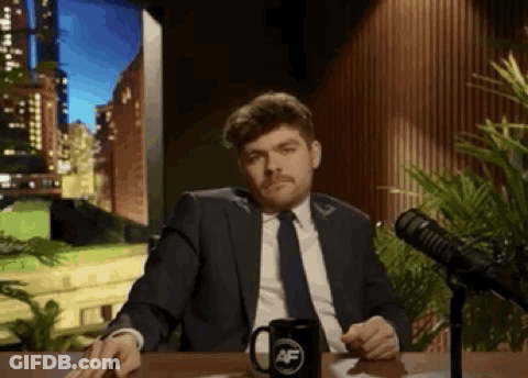 A Man In A Suit And Tie Is Sitting At A Table In Front Of A Microphone With A Mug On It . GIF