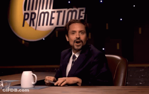 A Man In A Suit And Tie Is Sitting At A Table In Front Of A Sign That Says Primetime GIF