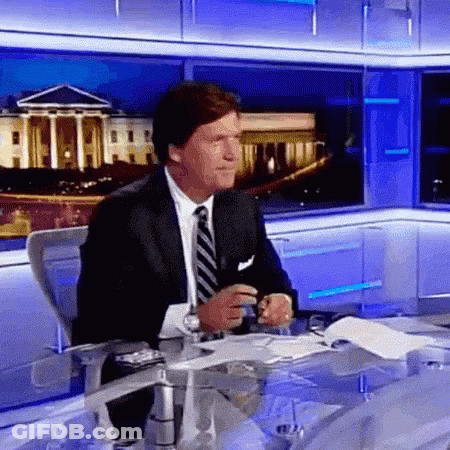 A Man In A Suit And Tie Is Sitting At A Table In Front Of A Television Screen . GIF