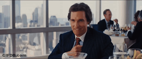 A Man In A Suit And Tie Is Sitting At A Table In Front Of A Window GIF