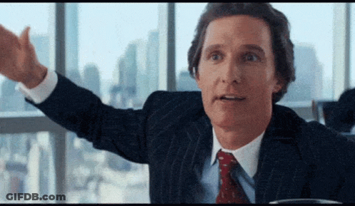A Man In A Suit And Tie Is Sitting At A Table In Front Of A Window With His Arms Outstretched . GIF