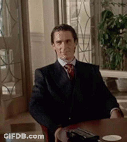 A Man In A Suit And Tie Is Sitting At A Table Looking At The Camera GIF