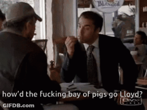 A Man In A Suit And Tie Is Sitting At A Table Talking To Another Man In A Hat . GIF